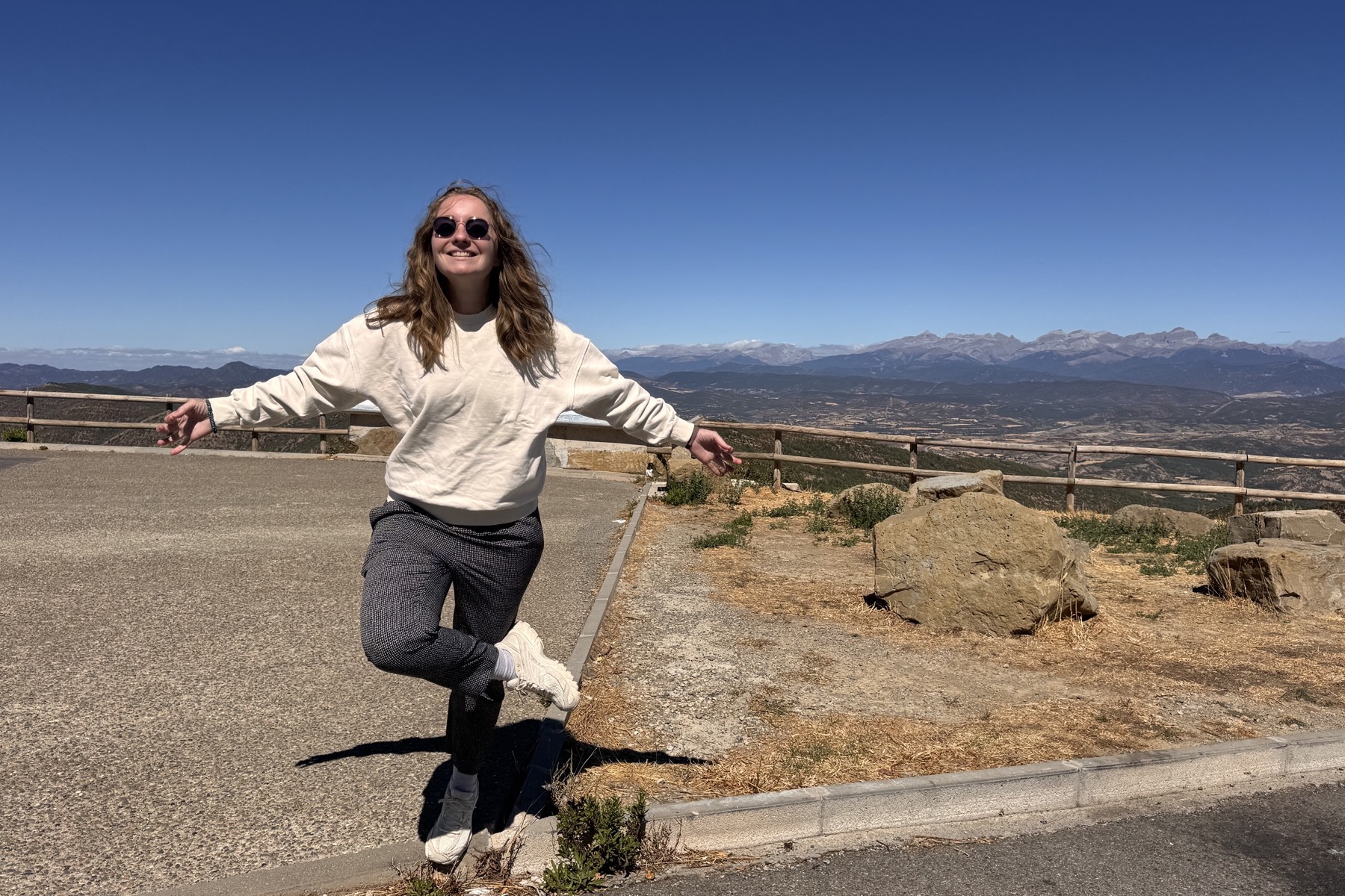 Charlotte Hibert, arms open, at a mountain viewpoint with the Pyrenees behind